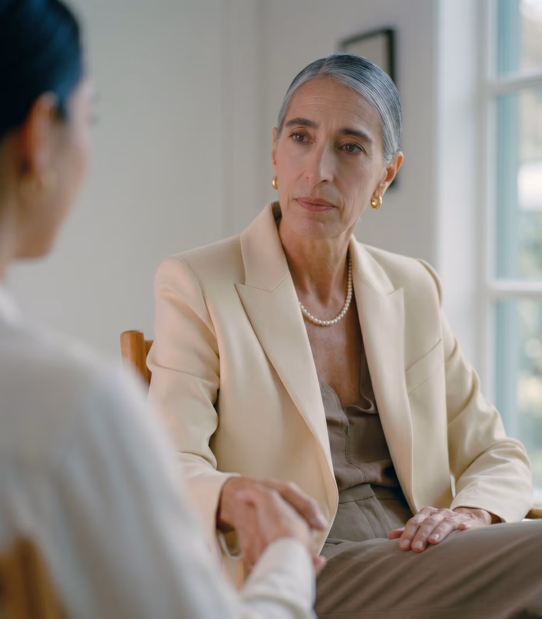 Serious middle-aged woman in beige blazer holding hands with another person in a calm setting.