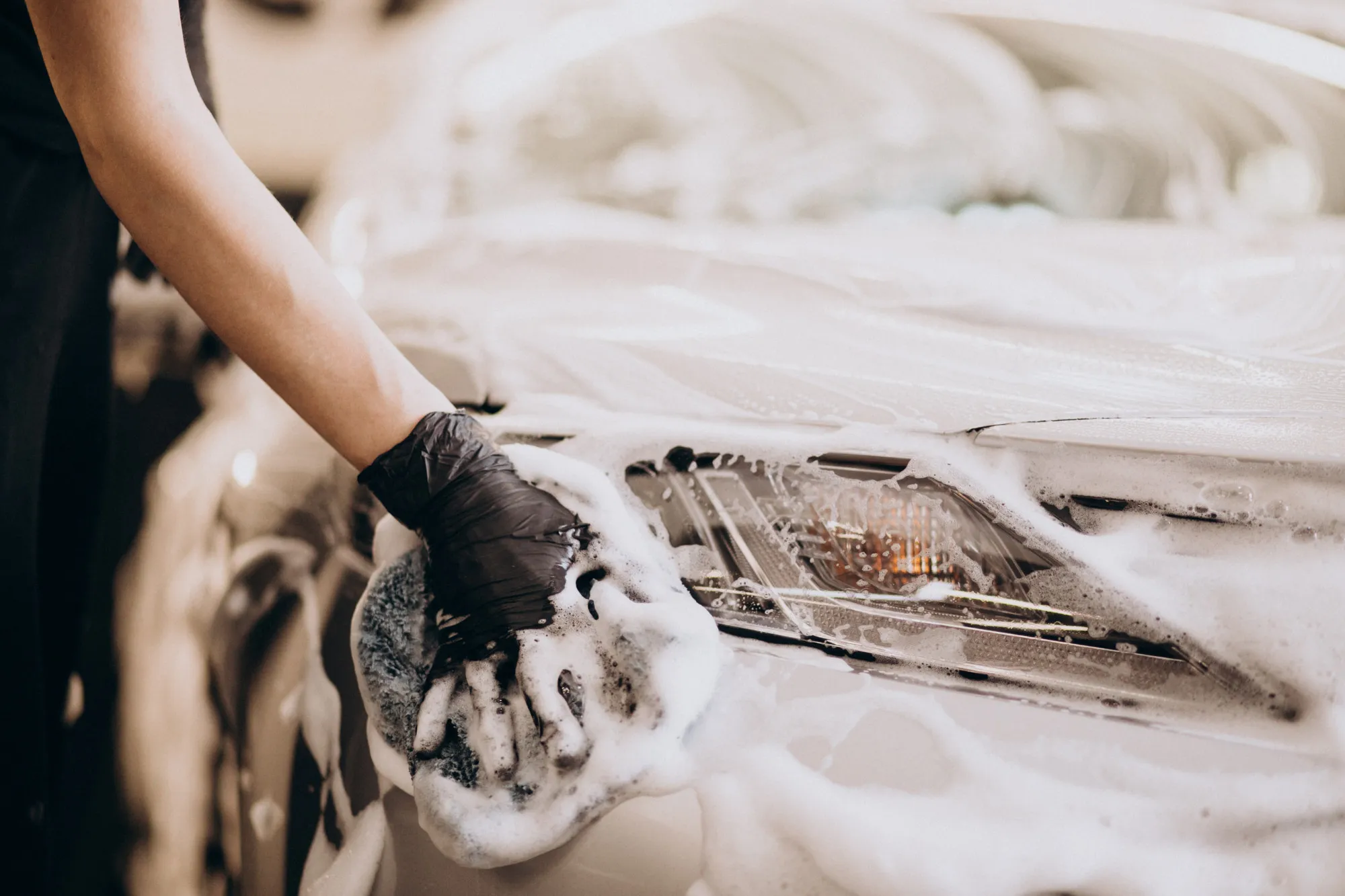 Person wearing black glove washing a car headlight with a soapy sponge.