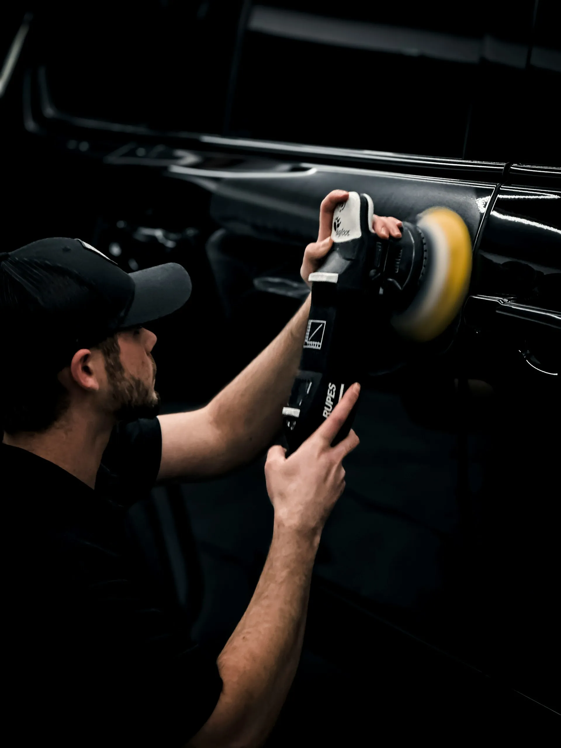 Man wearing a cap polishing a black car door with a handheld rotary buffer.
