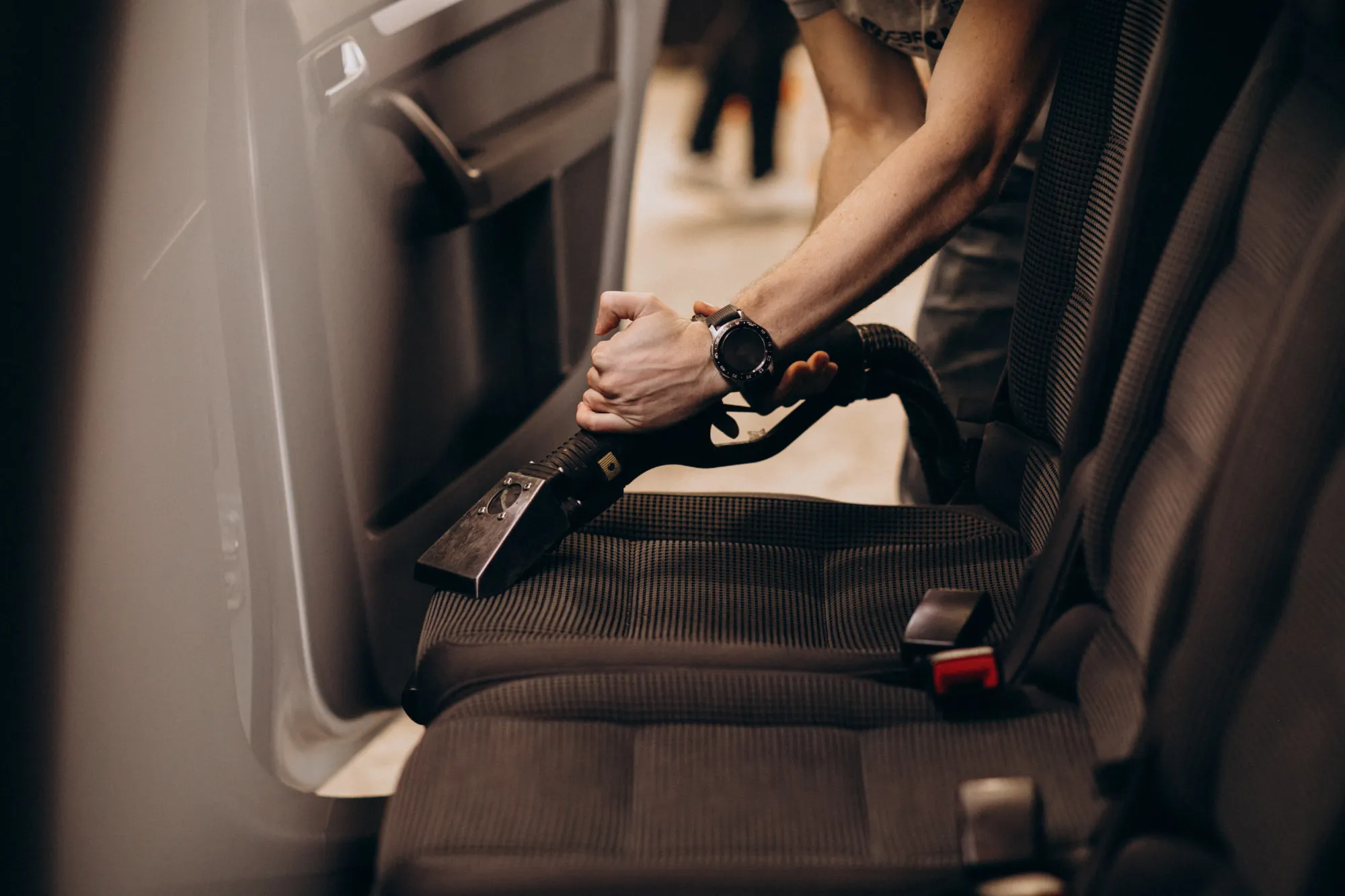 Person vacuuming the fabric seat inside a car with a handheld car vacuum cleaner.