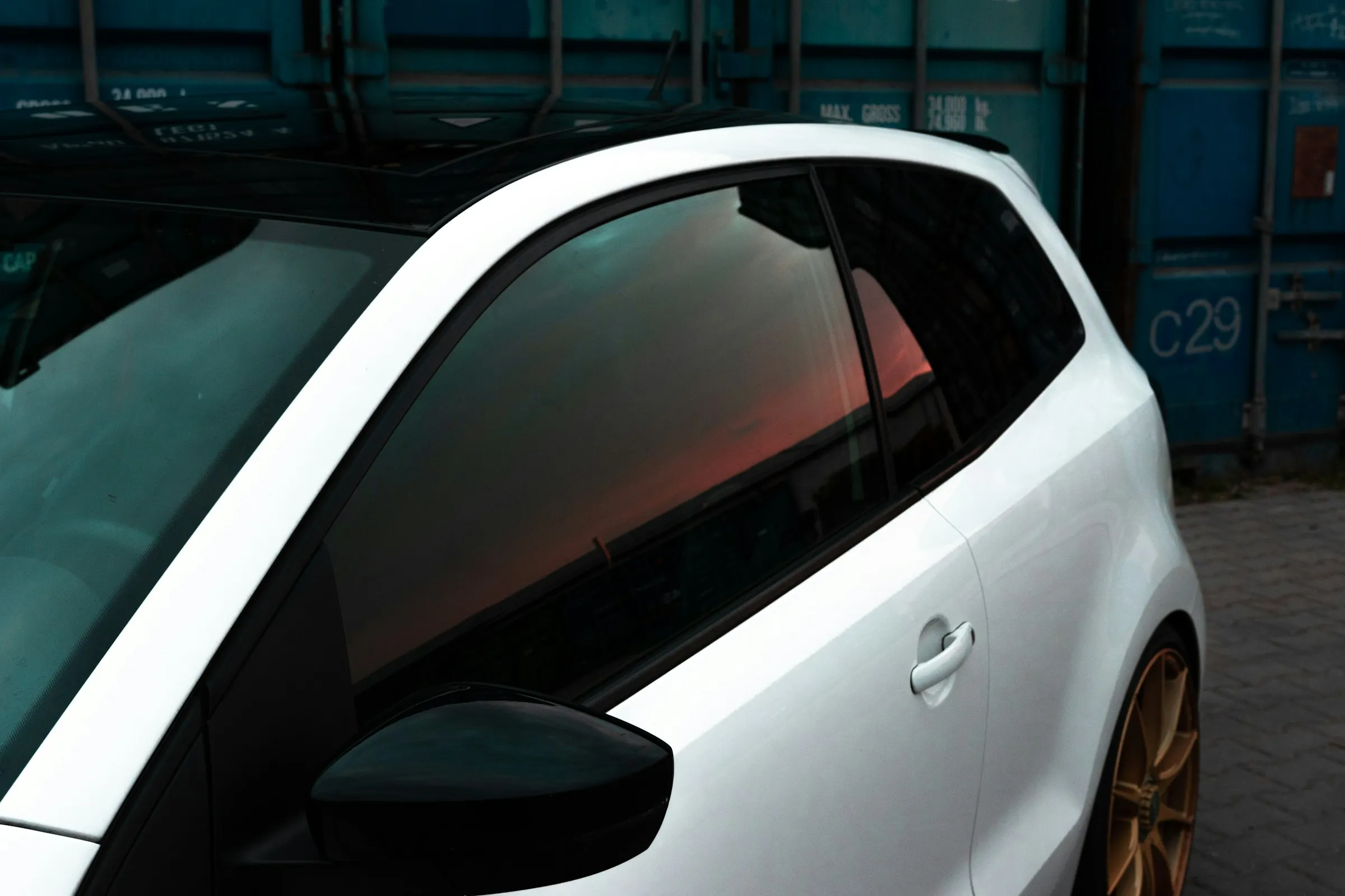 White car side with tinted windows reflecting a sunset and gold alloy wheels parked near blue containers.
