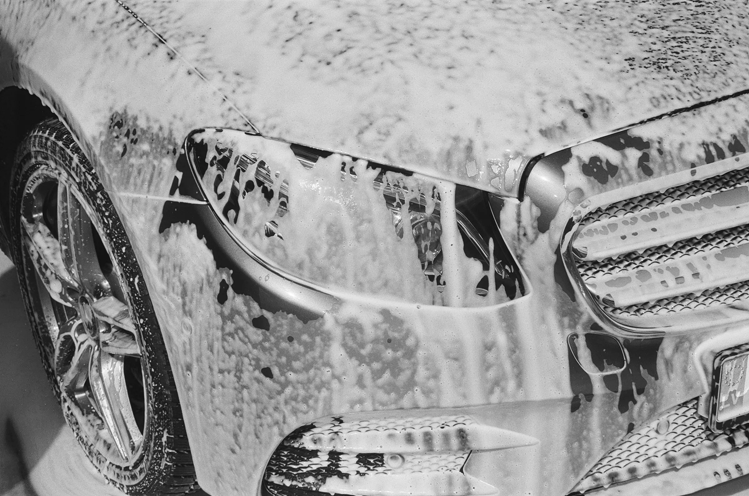 Close-up of a car covered in soap suds during a wash, showing the front wheel and headlight.