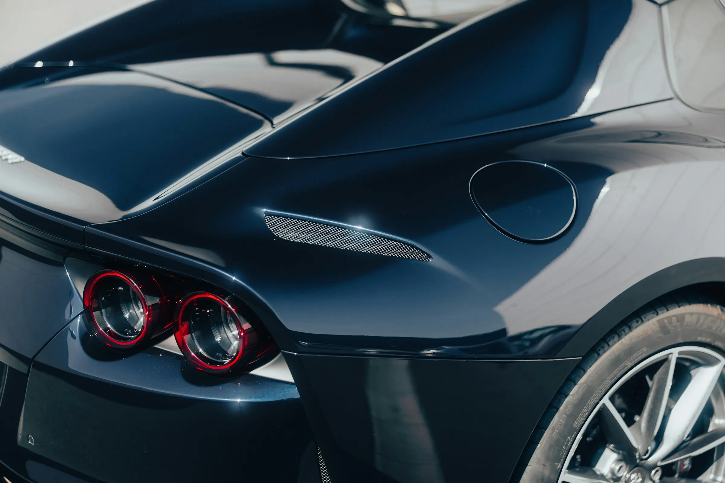 Close-up of rear side of dark blue sports car showing tail lights, fuel cap, and rear wheel.