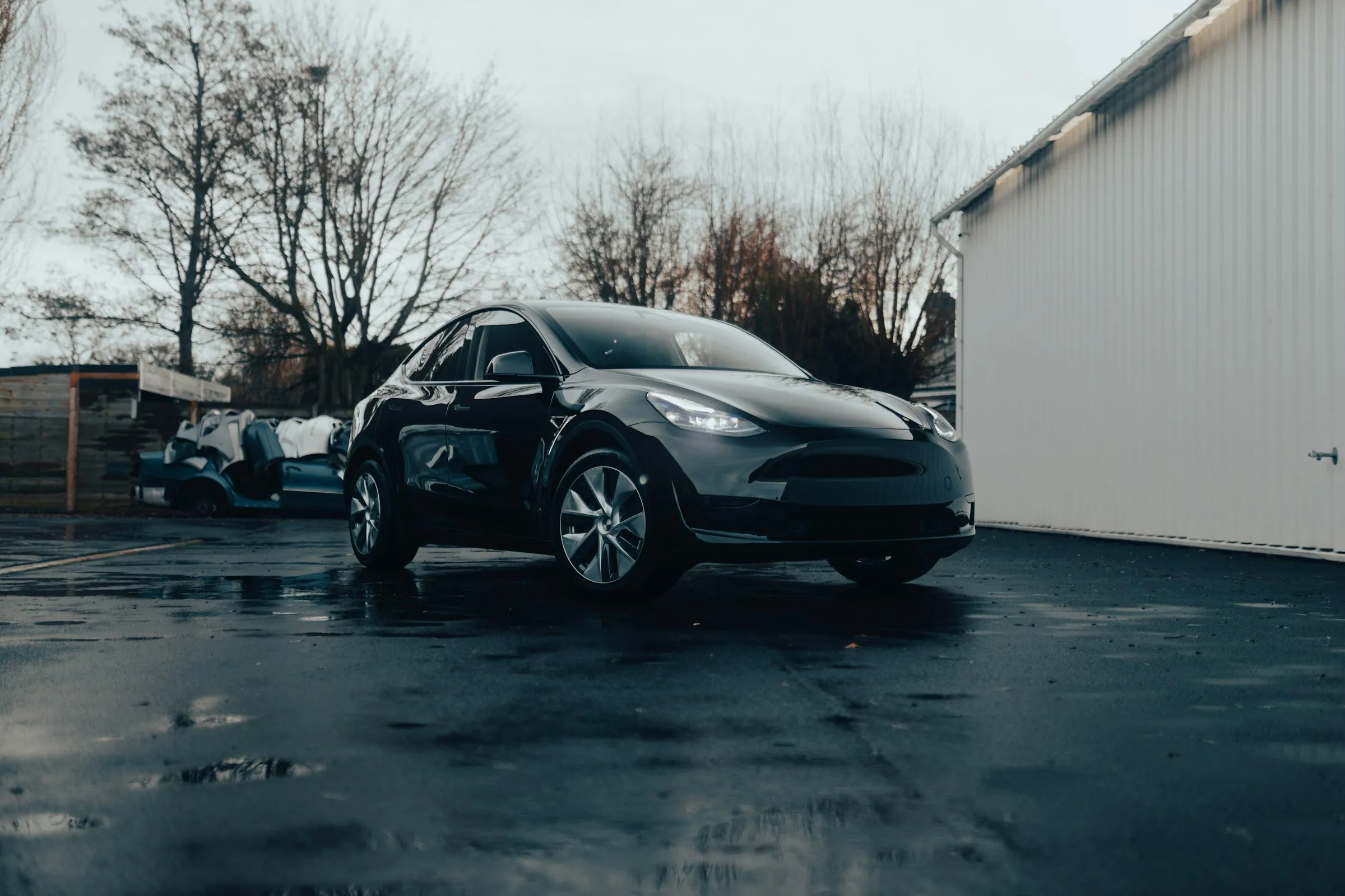 Black Tesla Model Y parked on wet pavement near a white building and leafless trees.