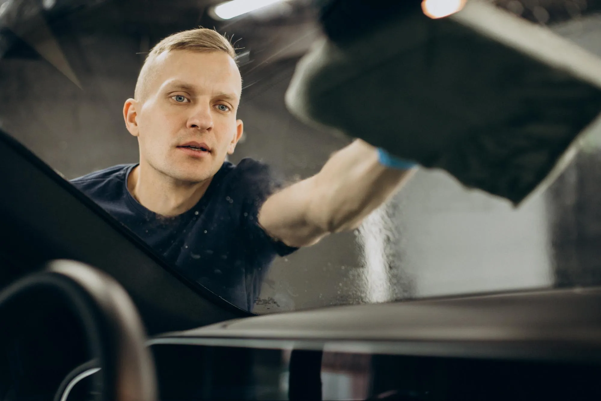 Man cleaning car windshield with a cloth inside a garage or workshop.