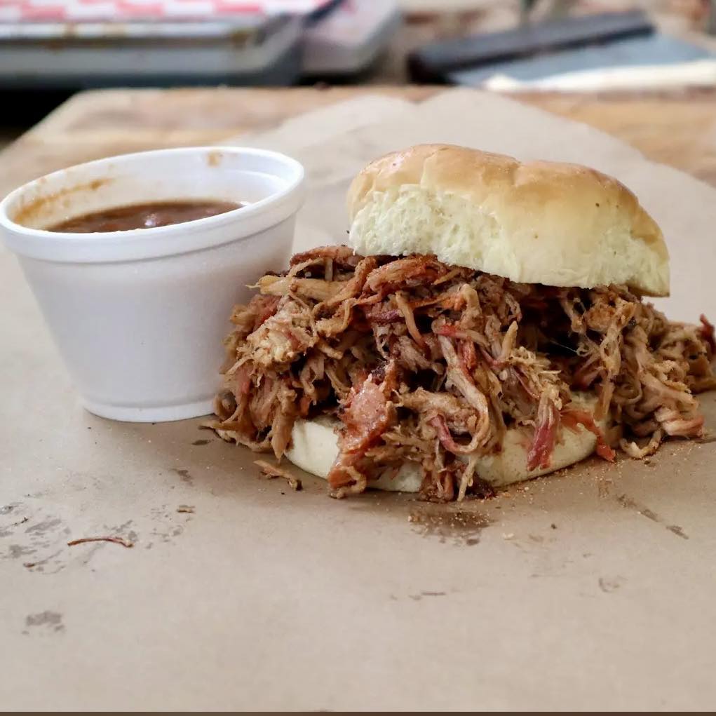 A sandwich with barbecue sauce and a cup of sauce on a paper plate.