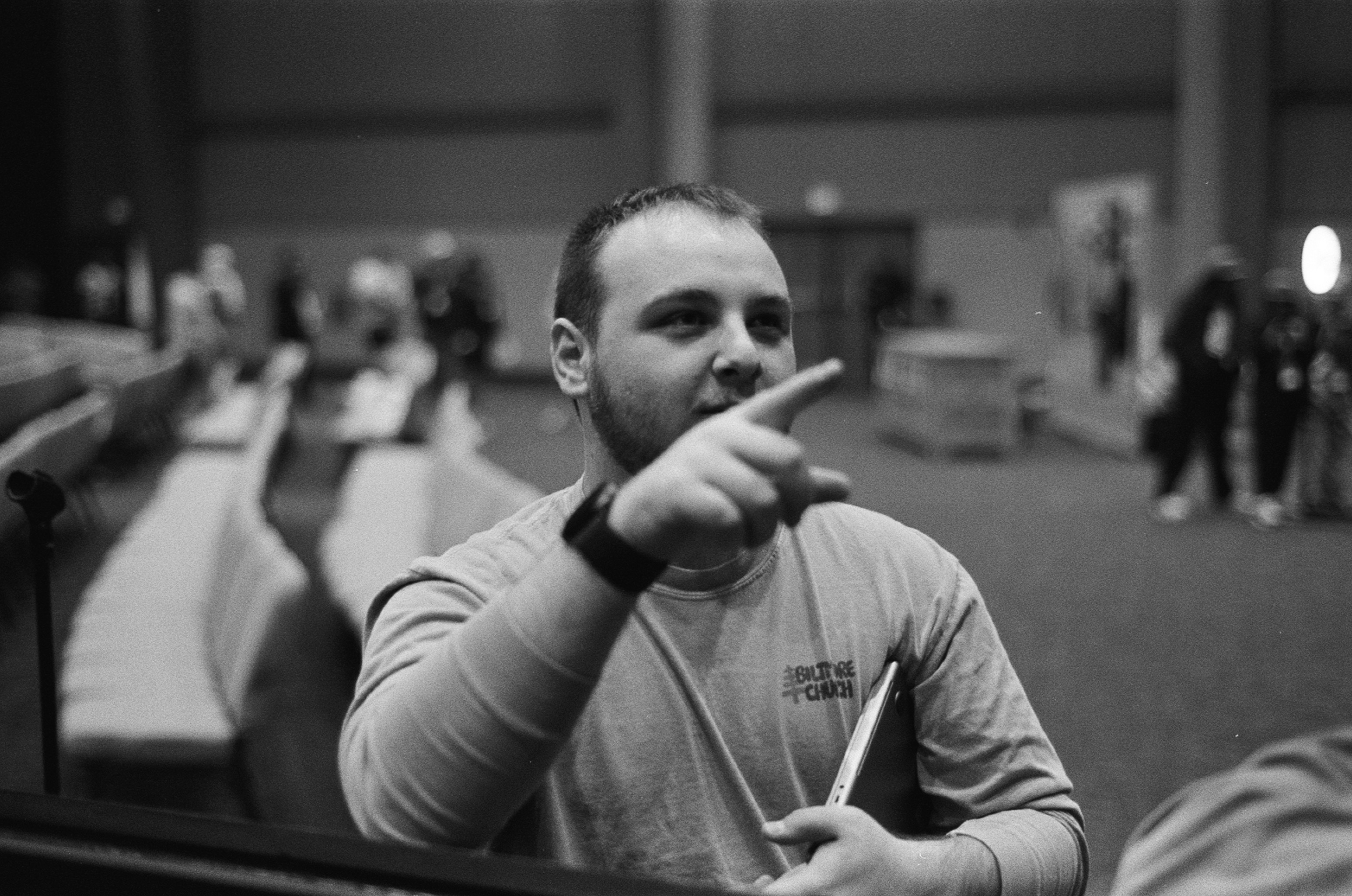 Man in a church shirt pointing forward while holding a laptop in a large room with chairs.