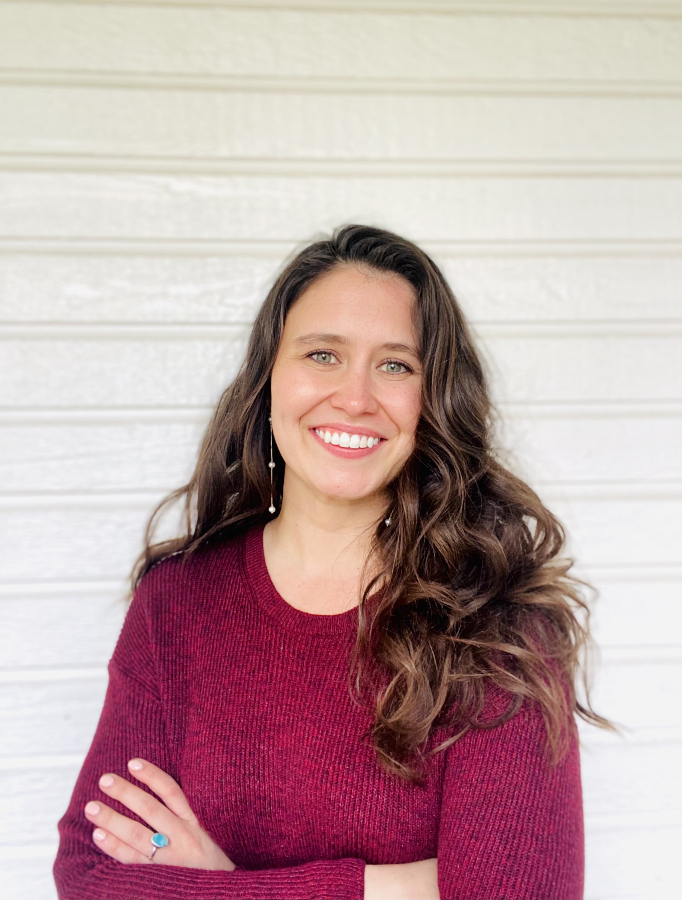 Smiling woman with long wavy brown hair wearing a burgundy sweater against a white paneled wall.