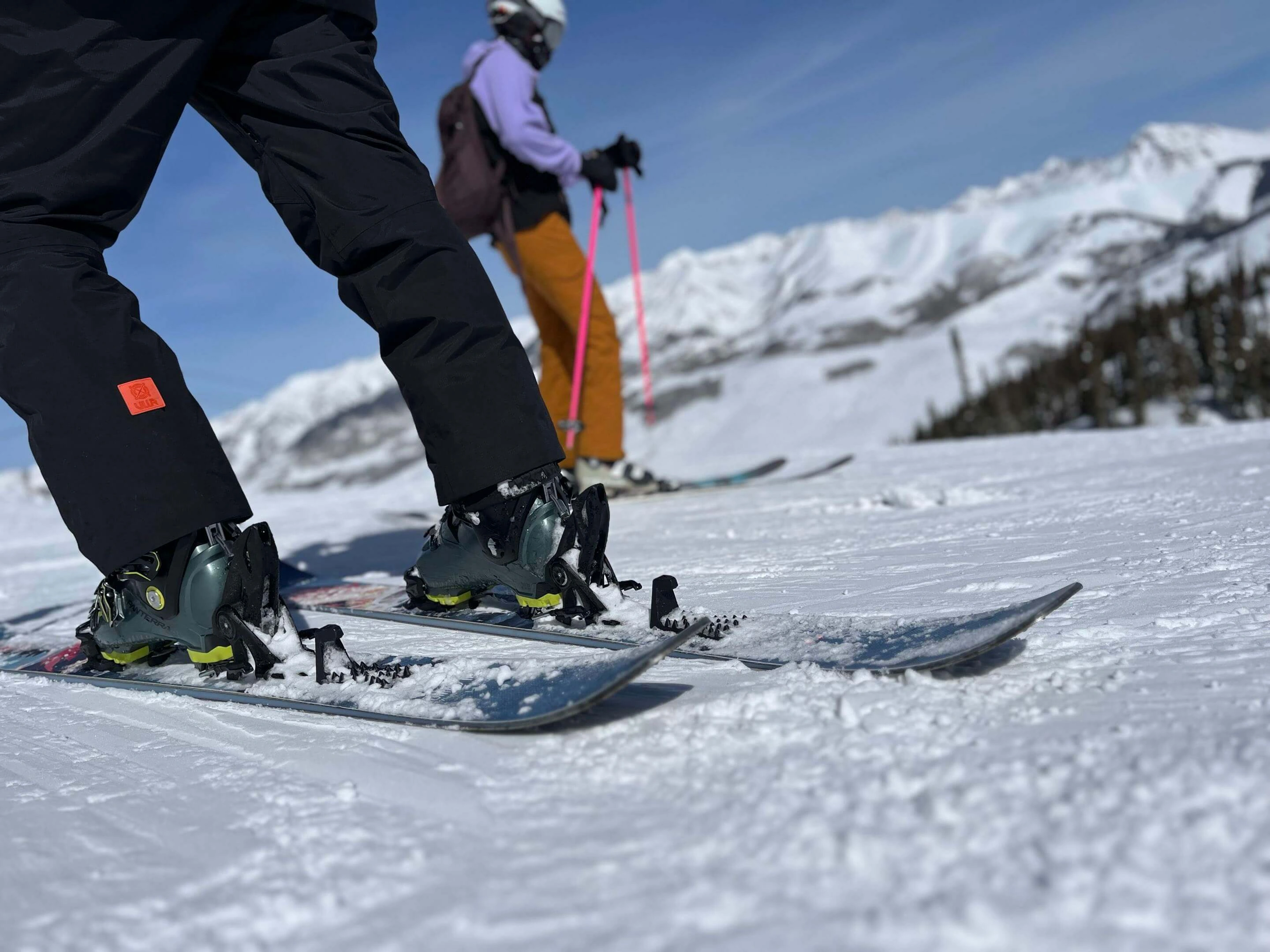 Close-up of a skier's boots and touring bindings on snow, another skier visible in the background on a sunny mountain.