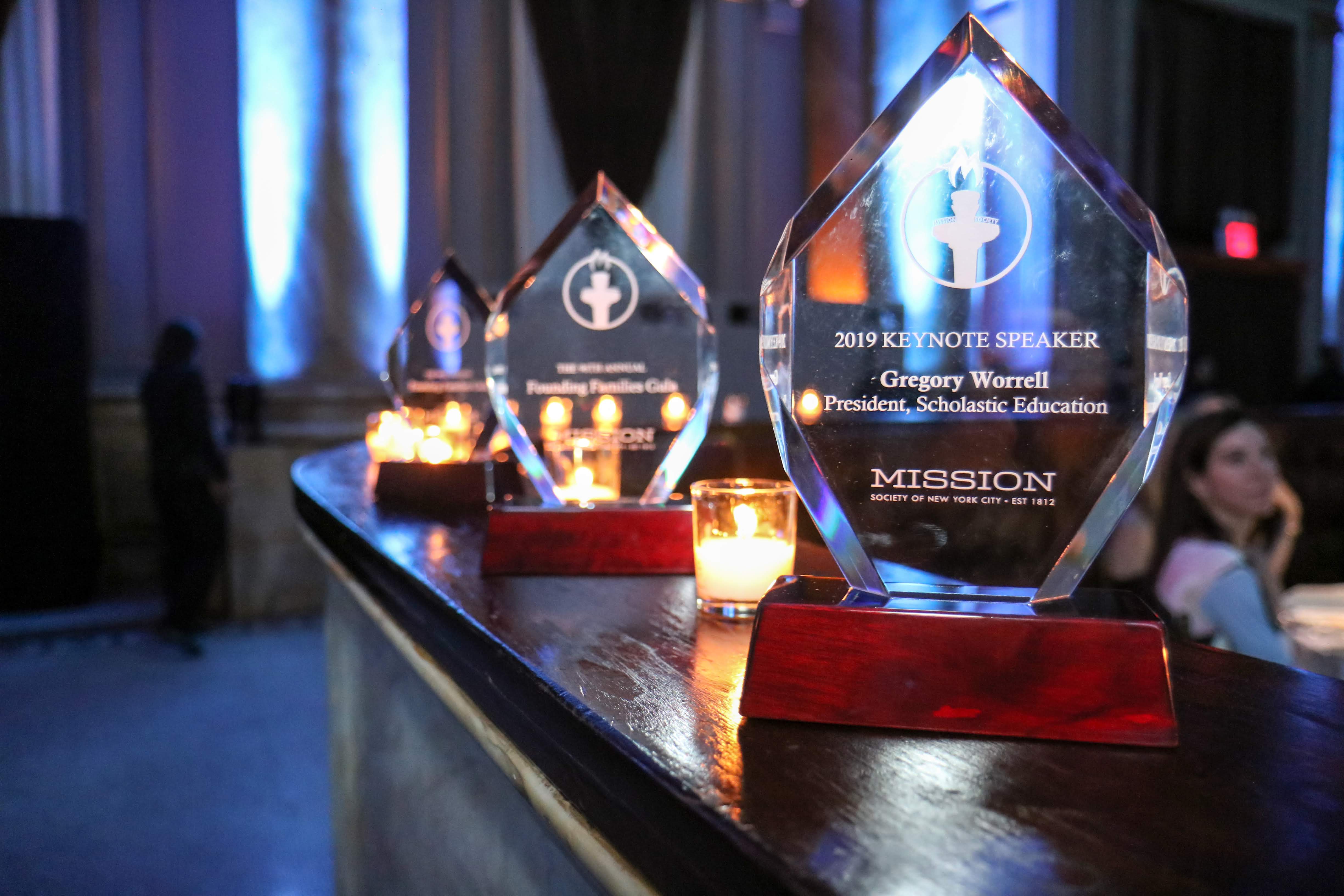 Row of clear glass awards and lit candles on a dark wooden surface at an event.