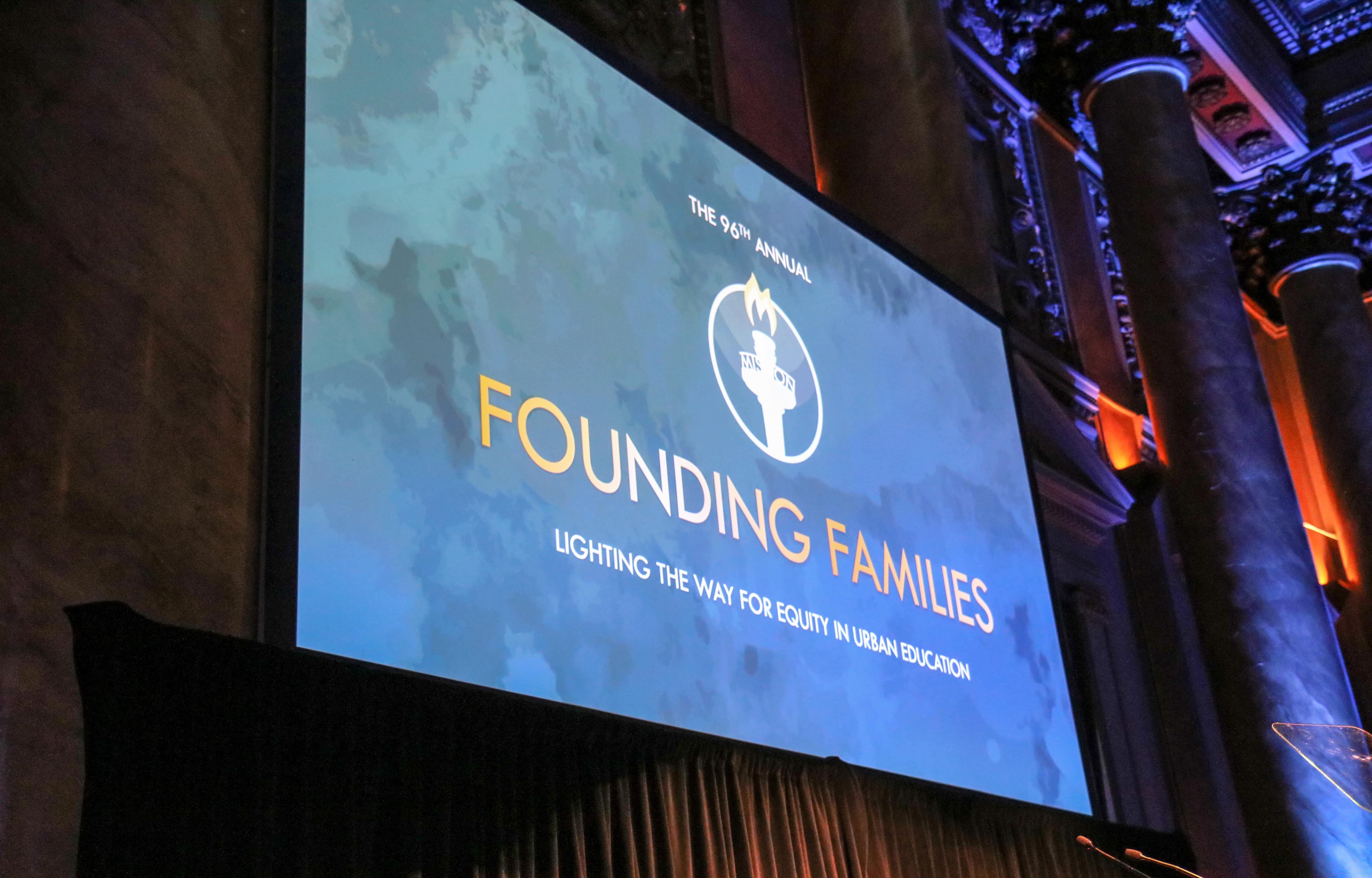 Large screen displaying text 'The 96th Annual Founding Families' with a torch logo in a grand hall.