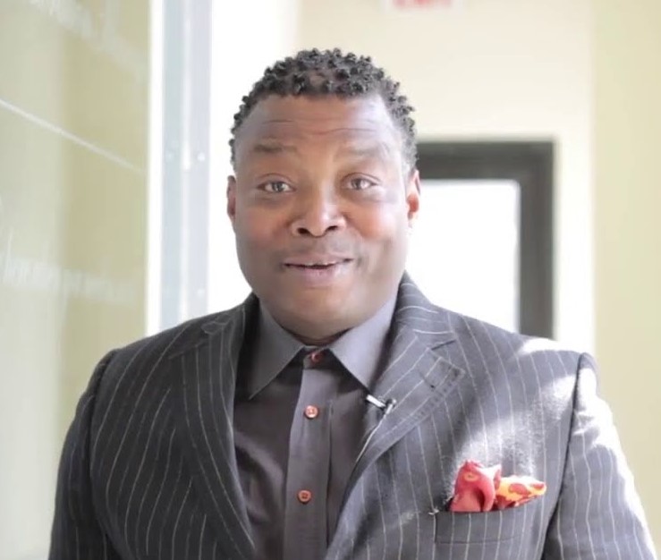 Man with short curly hair wearing a dark pinstripe suit and red pocket square indoors.