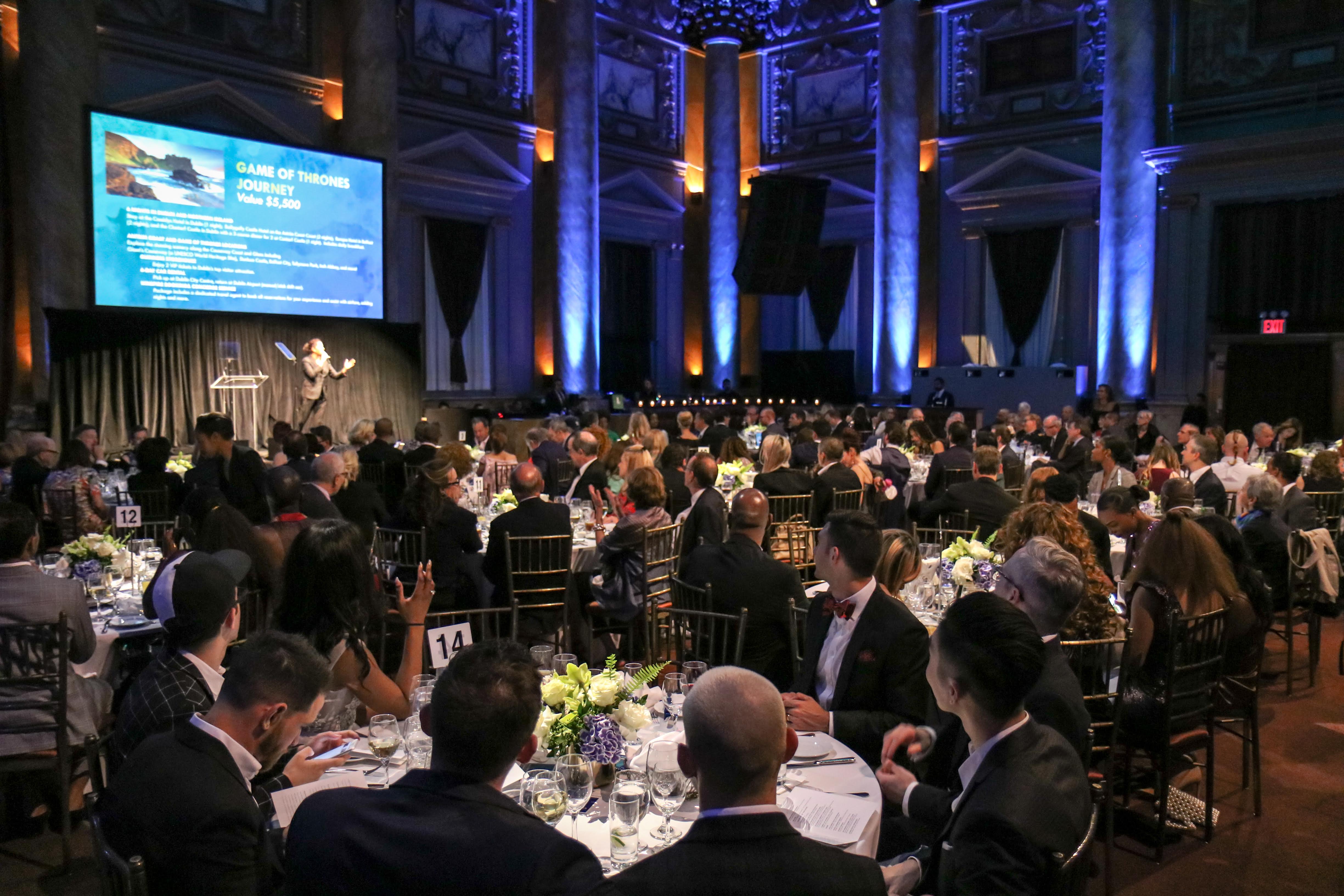 Large group seated at round tables in formal event hall with blue lighting and speaker on stage.