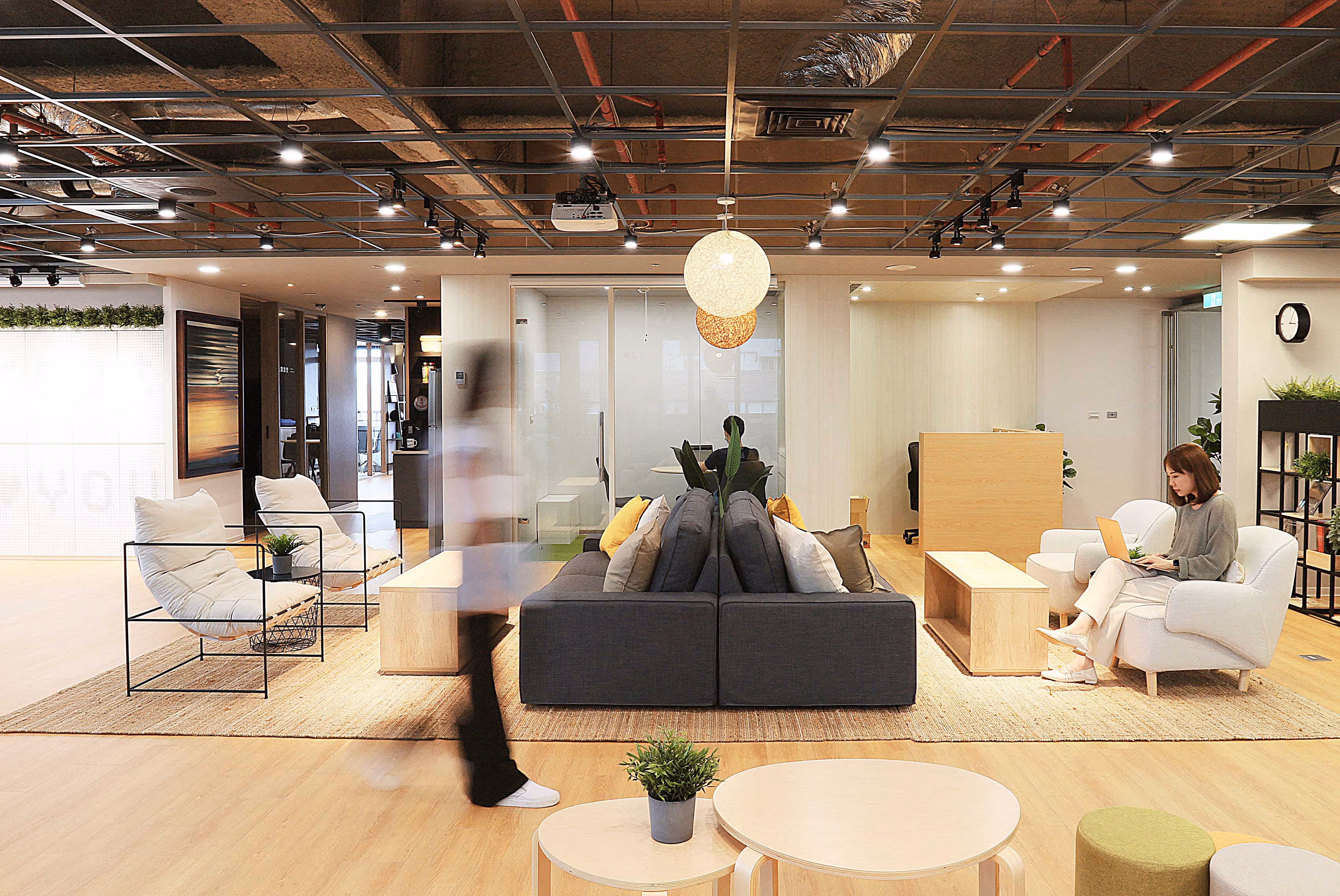 Modern office lounge with a woman using a laptop, cozy seating, wooden tables, and hanging lights.