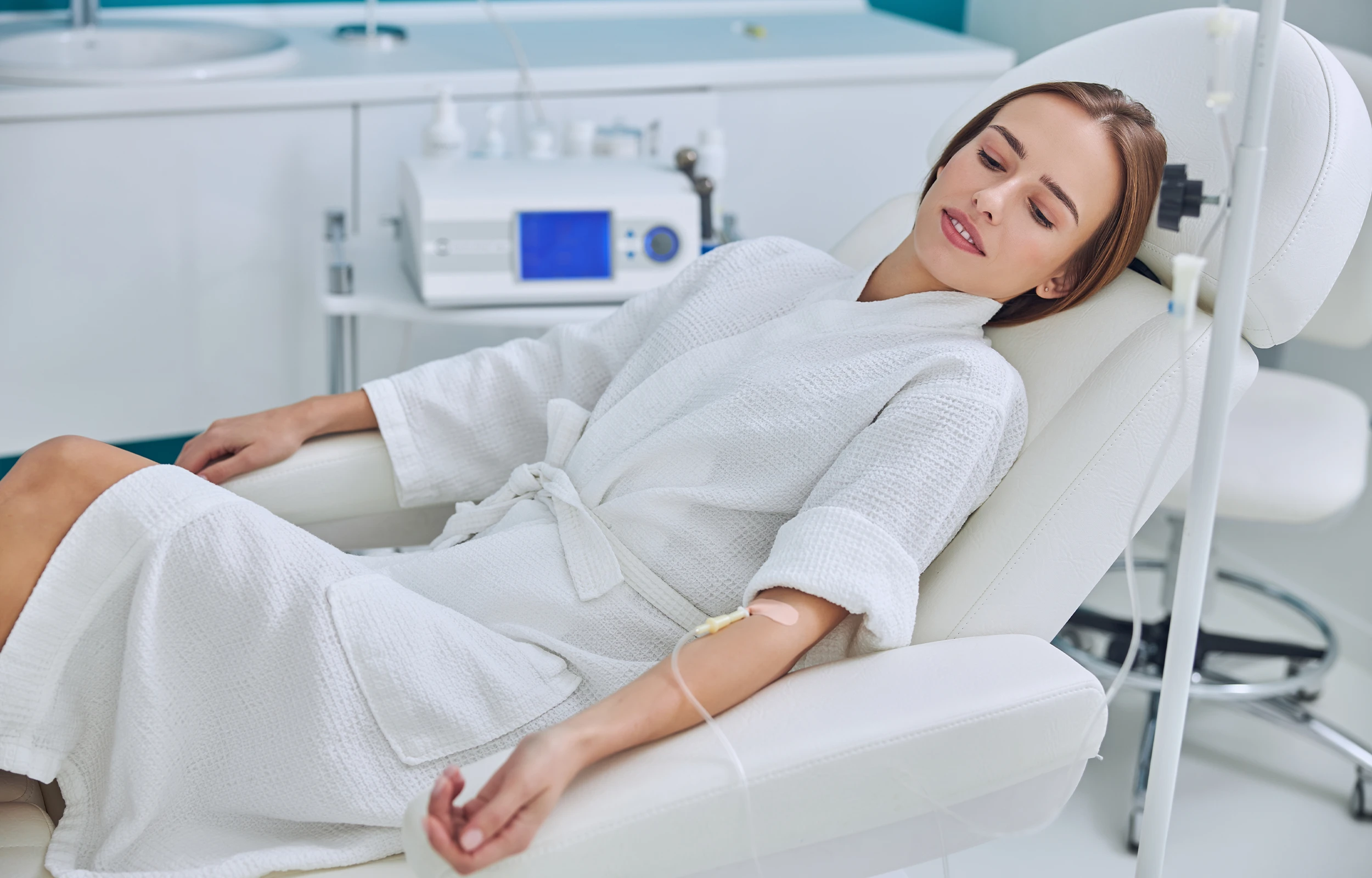 Woman in white robe reclining on a medical chair receiving intravenous therapy in a clinical setting.