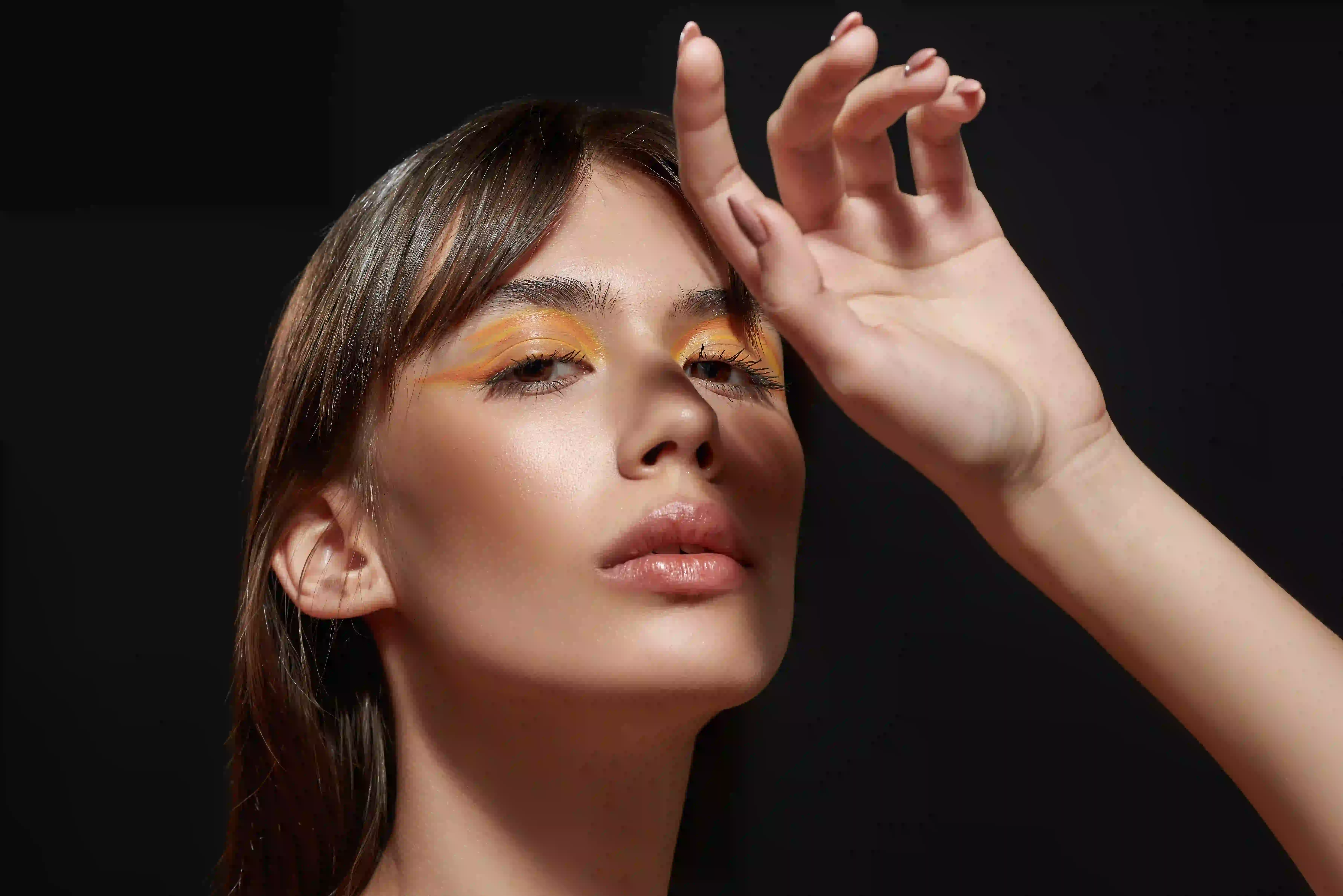 Close-up of a woman with yellow eyeshadow raising her hand against a dark background.