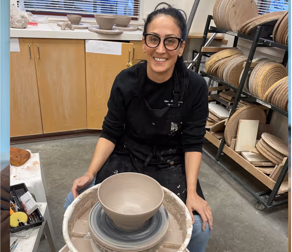 Smiling woman shaping a clay bowl on a pottery wheel in a ceramics studio.