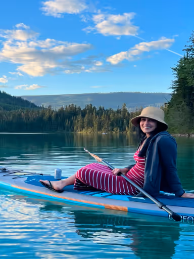 Smiling woman in a striped dress and hat sitting on paddleboard on calm lake surrounded by trees.