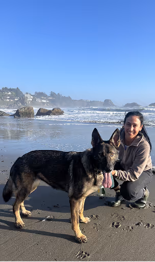 Woman smiling and crouching beside a large black and tan German shepherd on a sandy beach with waves.