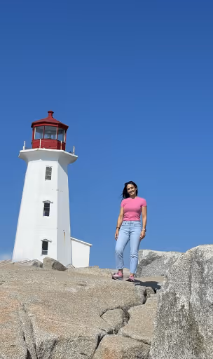 Woman in pink shirt and jeans standing on rocky ground near a white lighthouse under blue sky.