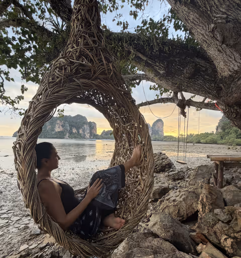 Woman sitting in a woven hanging chair by the rocky shore with sea and cliffs at sunset.