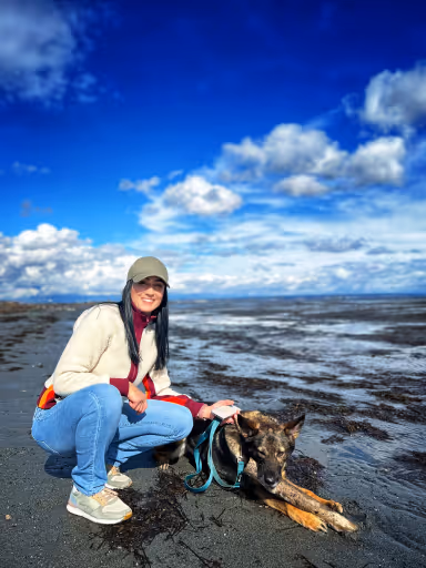 Woman in jeans and sweater crouching beside a German Shepherd on a sandy beach under a blue sky.