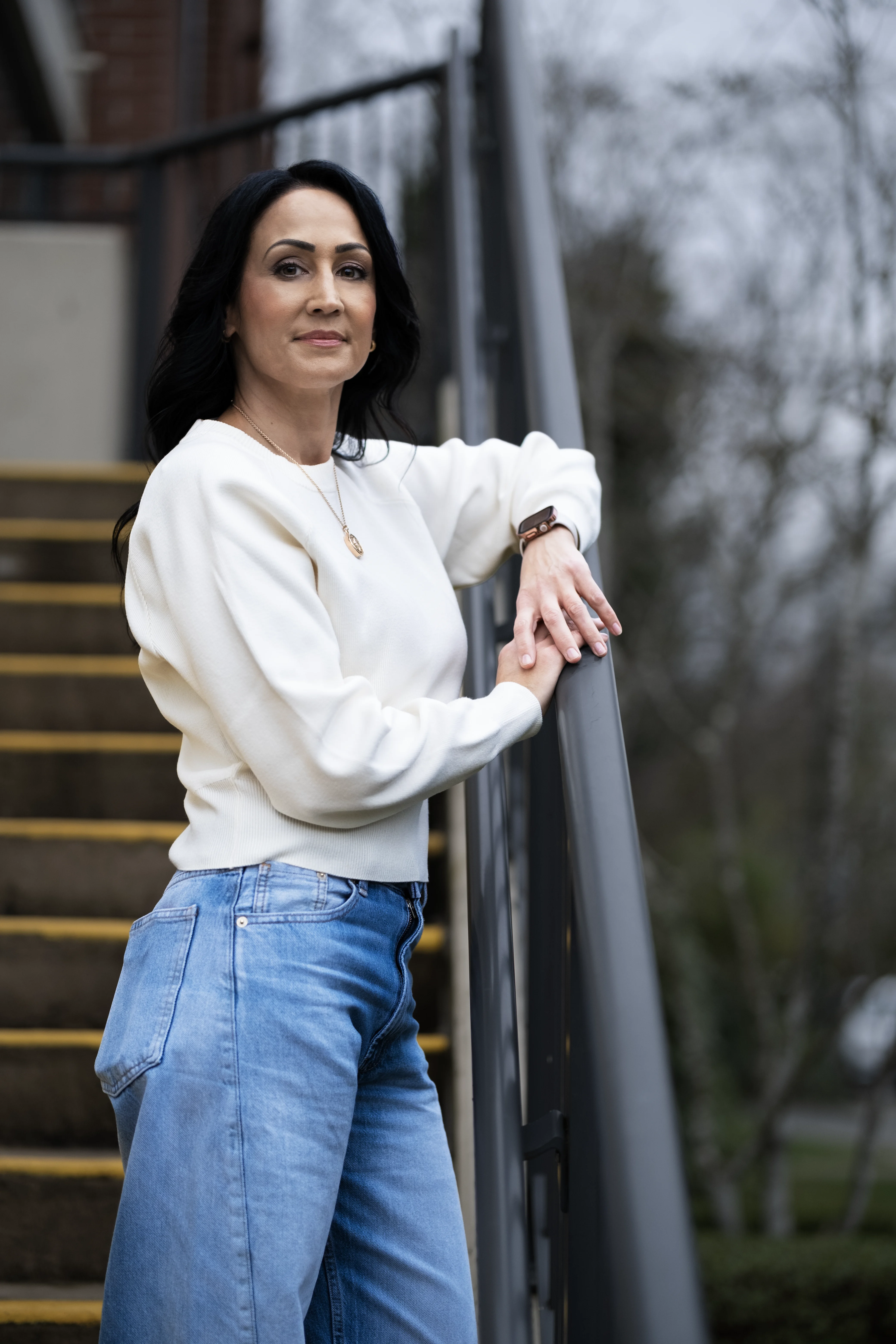 Woman in white sweater and blue jeans leaning on stair railing outdoors.
