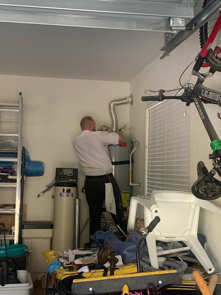 Technician working on a water heater in a cluttered garage with tools, ladders, and a bicycle.