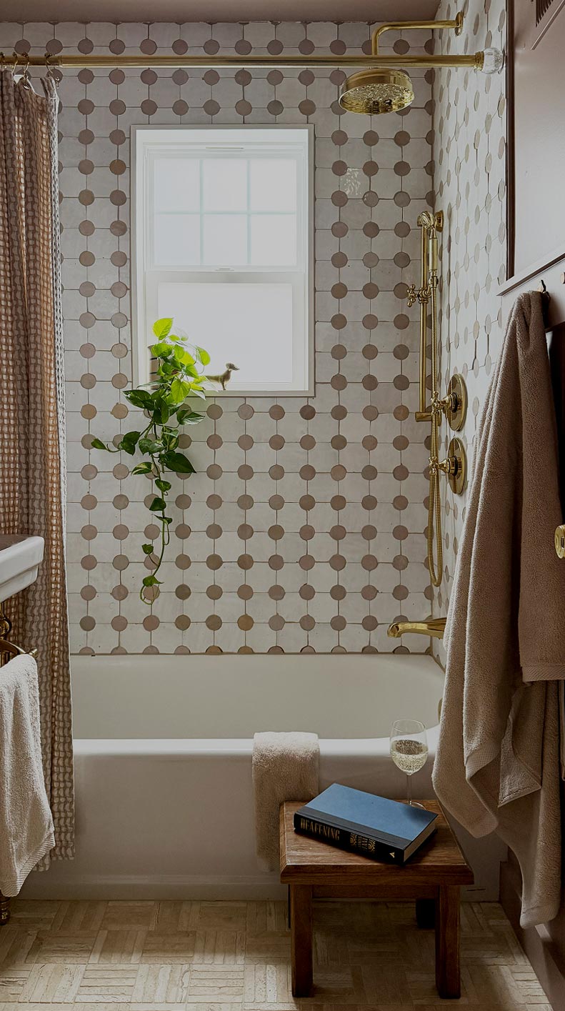 Bathroom bathtub with gold shower fixtures, patterned tile wall, hanging plant, book, and glass of wine.