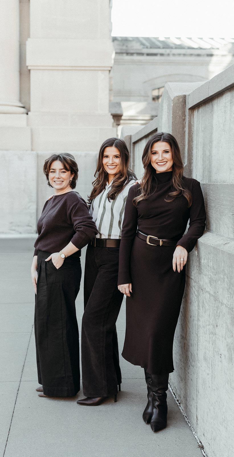 Three smiling women standing outdoors against a stone wall and pillars in stylish black and white outfits.