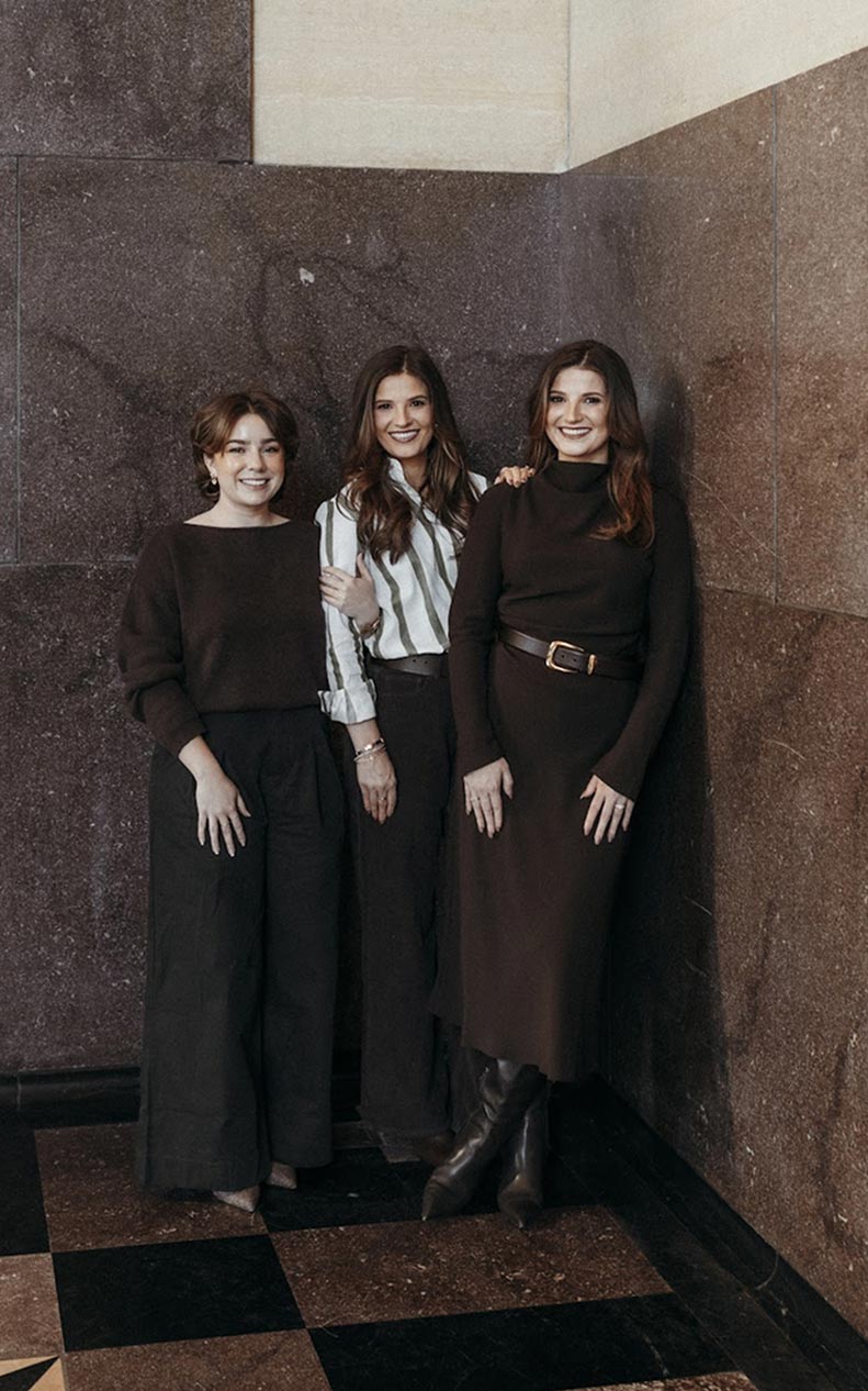 Three women standing indoors against a brown marble wall, smiling and posing for the photo.