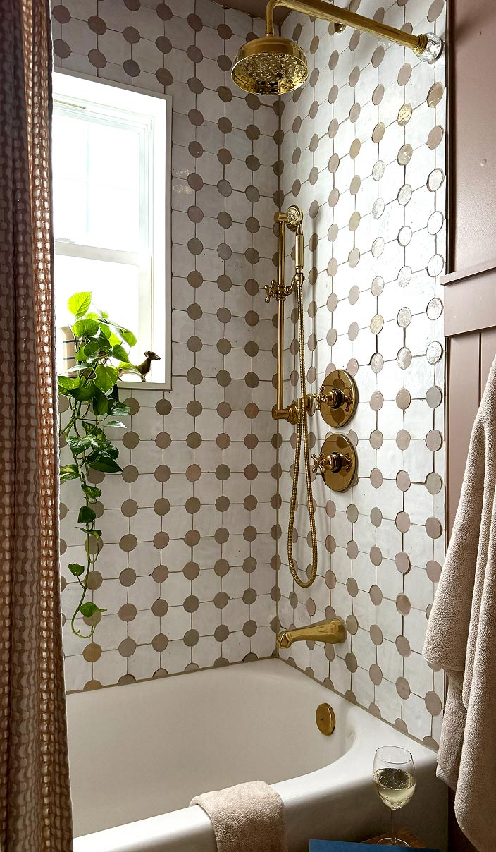 Bathroom with white bathtub, gold shower fixtures, patterned tile wall, plant by window, and glass of wine.