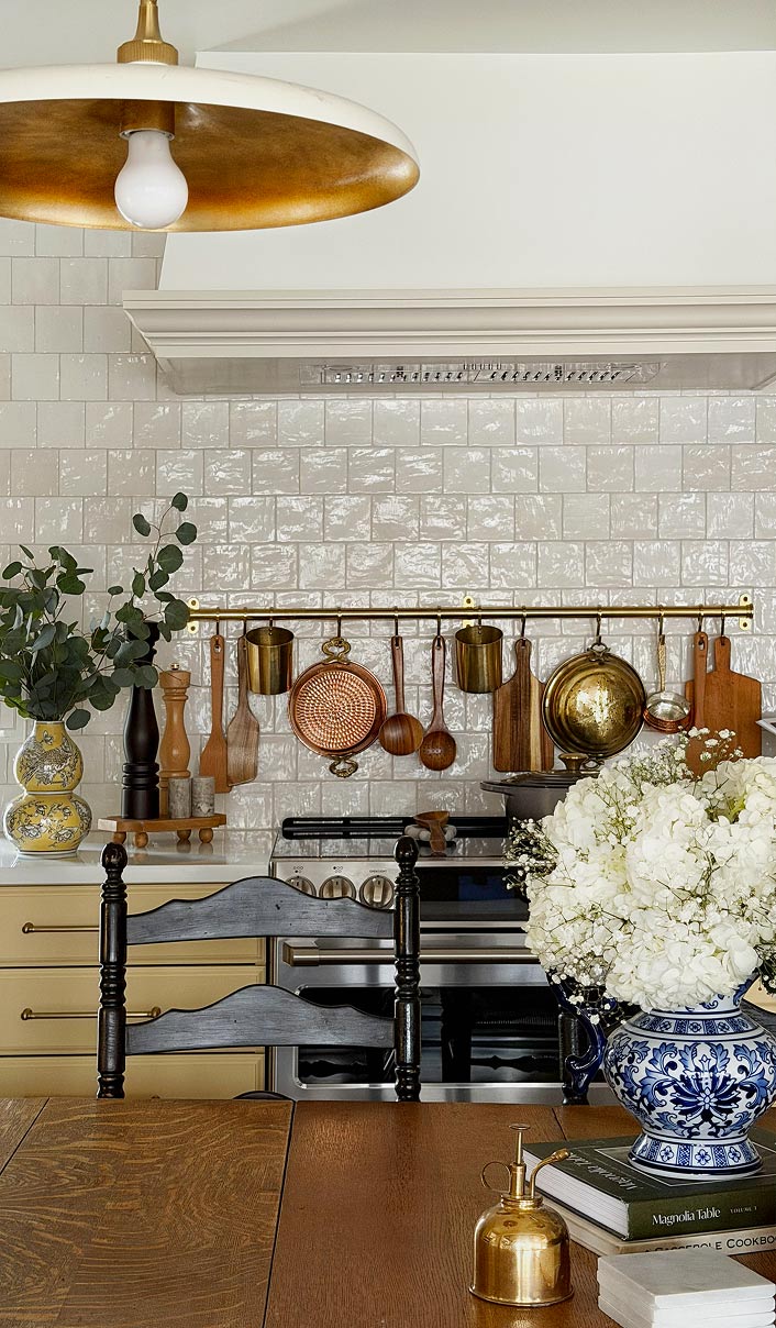 Cozy kitchen with hanging copper pans, wooden utensils, a stove, dark chair, and floral vase on wooden table.