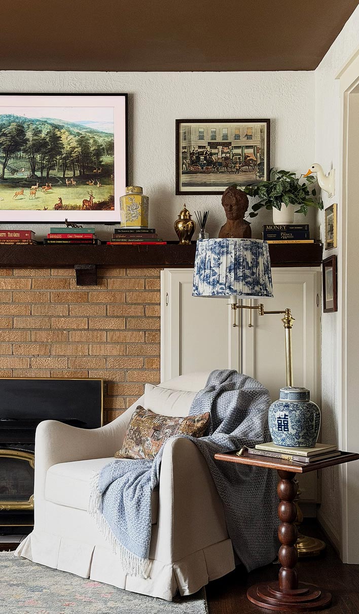 Cozy living room corner with cream armchair, blue throw, patterned pillow, side table, and wall art above mantel.