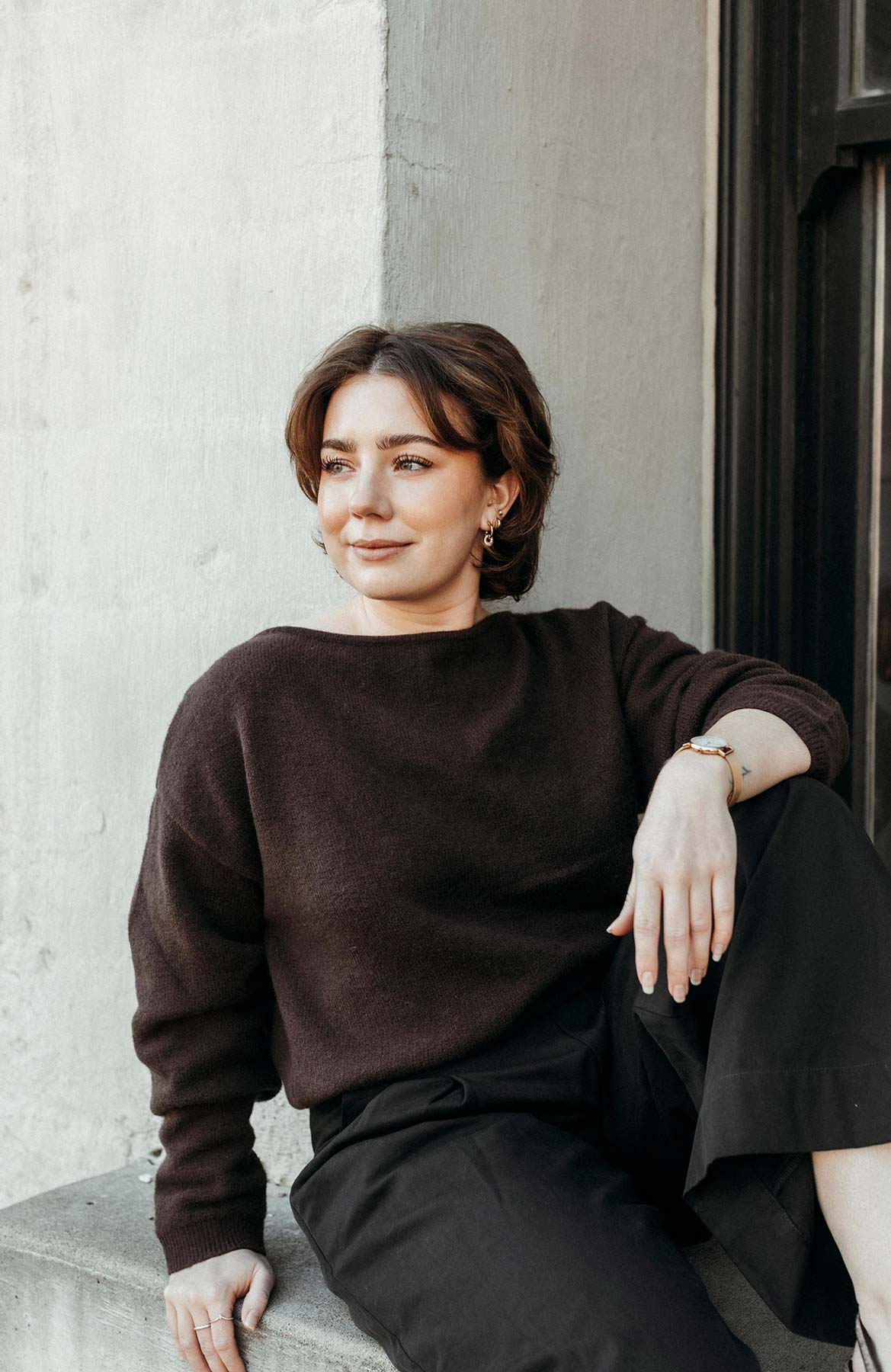 Young woman with short brown hair in dark sweater sitting on a concrete ledge, looking left.