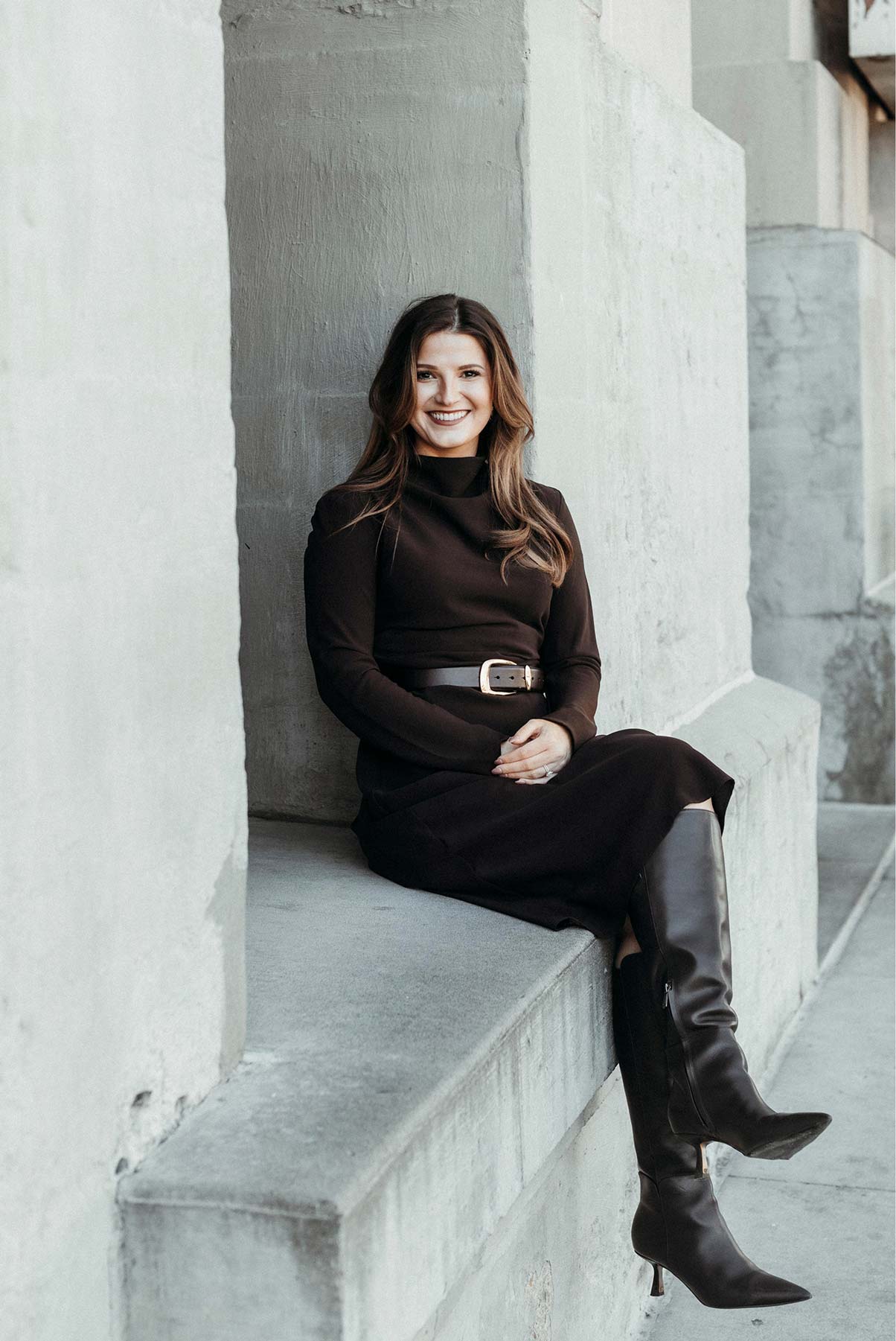 Woman in black dress and tall boots sitting on stone ledge, smiling with hands clasped.