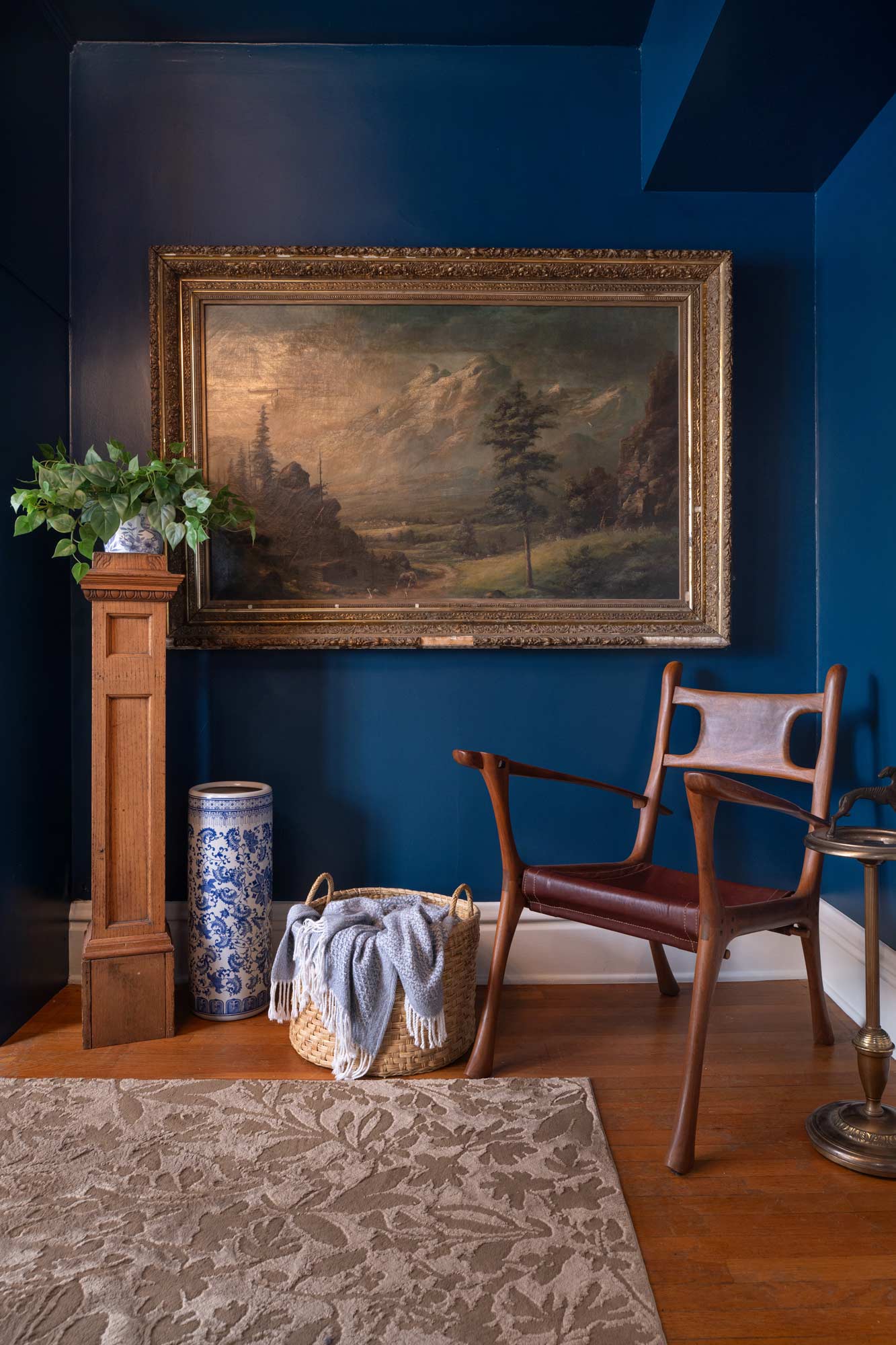 Wooden chair with leather seat near basket with blanket, plant on pedestal, and mountain landscape painting.