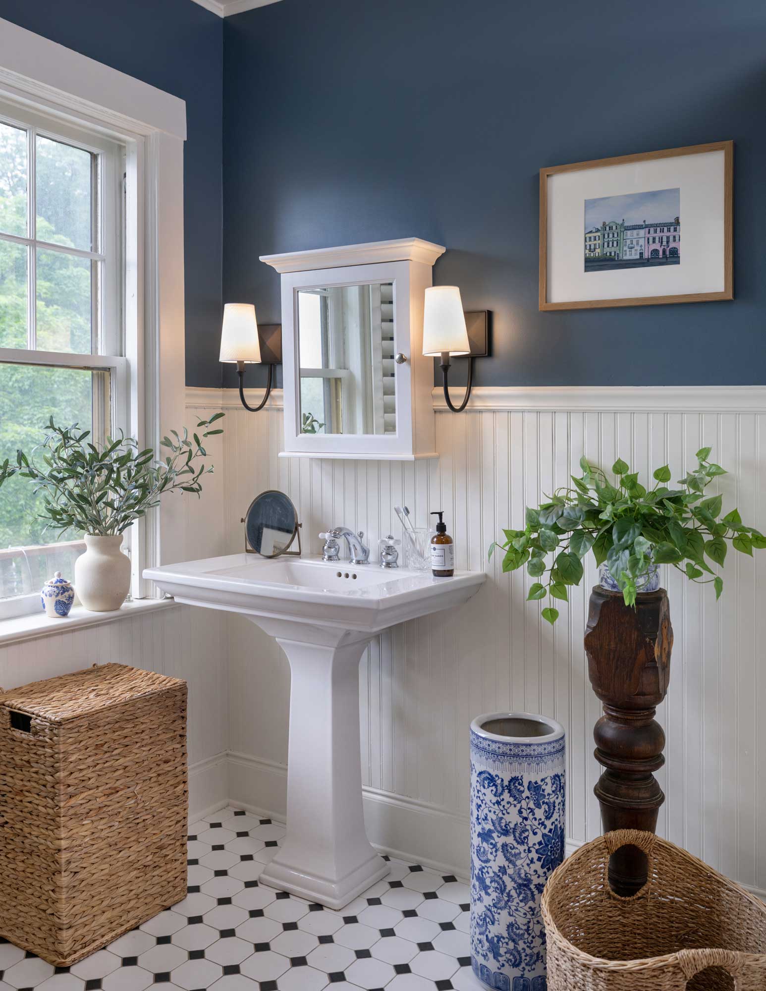 Bathroom with blue wall, white pedestal sink, wicker baskets, plants, and framed artwork.
