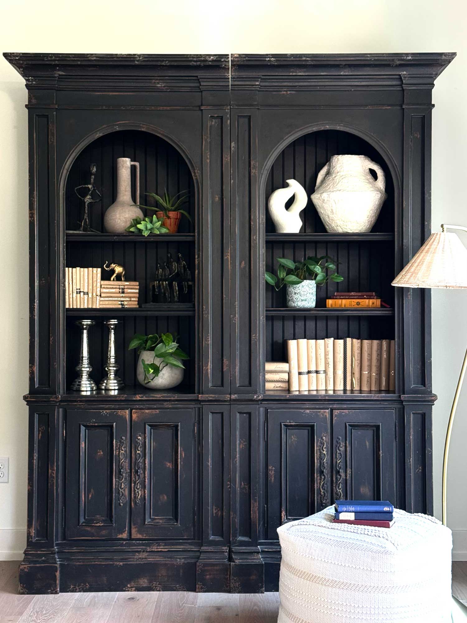 Black distressed wooden cabinet with shelves holding plants, books, vases, and decorative objects.