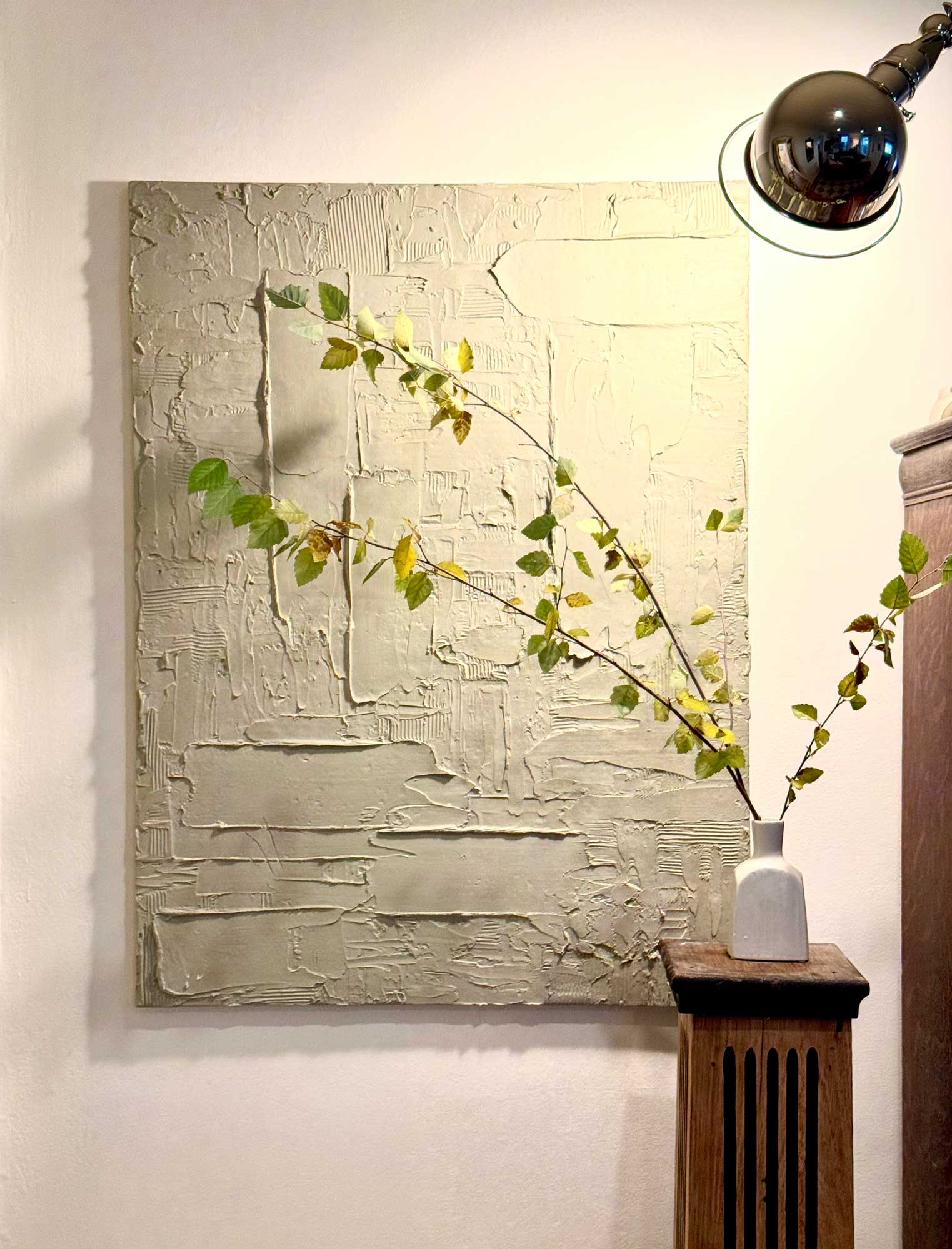 Minimalist interior with textured beige wall art, a tall plant in a white vase on wooden stand, and a black lamp.