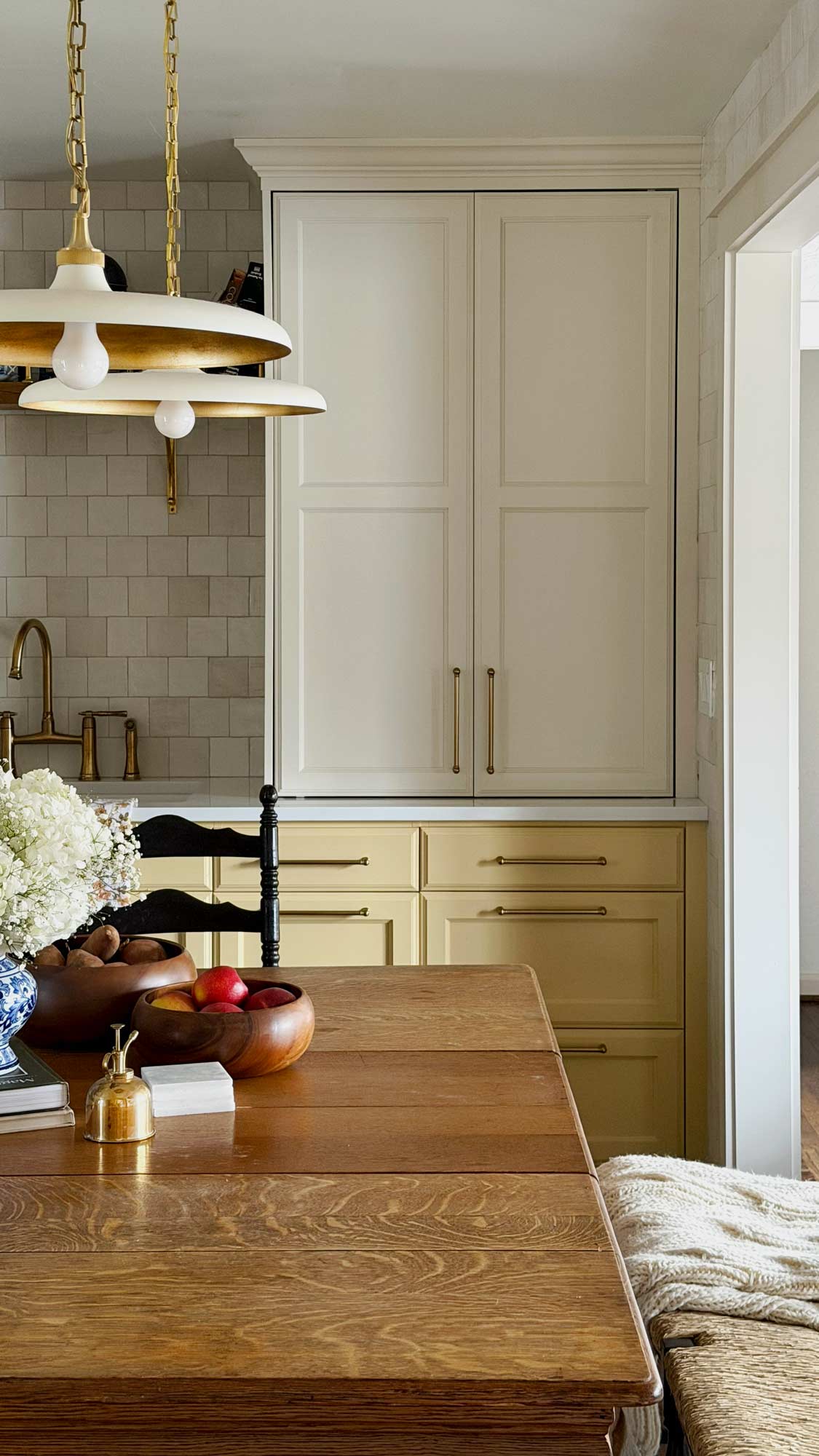 Wooden kitchen table with fruit bowls, flowers, brass tap, pendant lights, and cream and yellow cabinets.