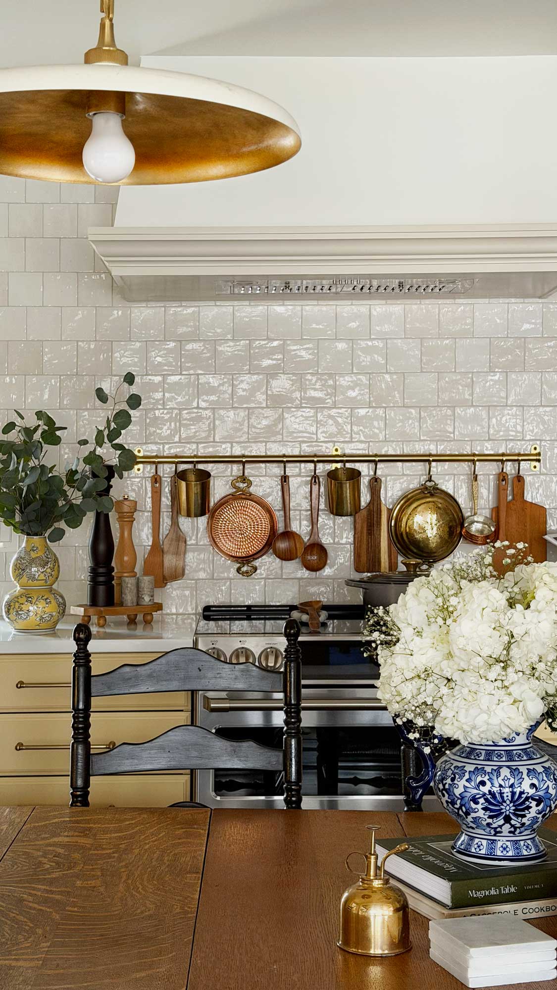 Cozy kitchen with hanging copper pots, wooden utensils, floral vases, and a wooden dining table.