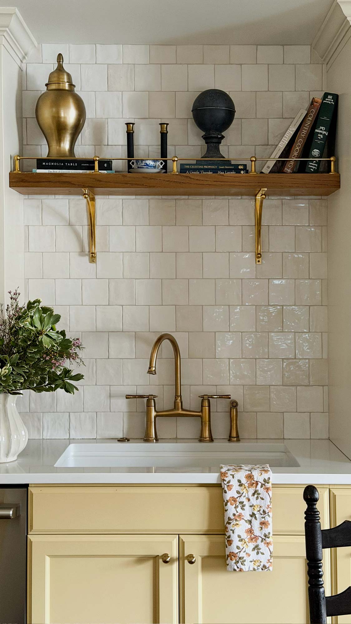 Kitchen sink with brass faucet below wooden shelf holding decor and books against tiled wall.