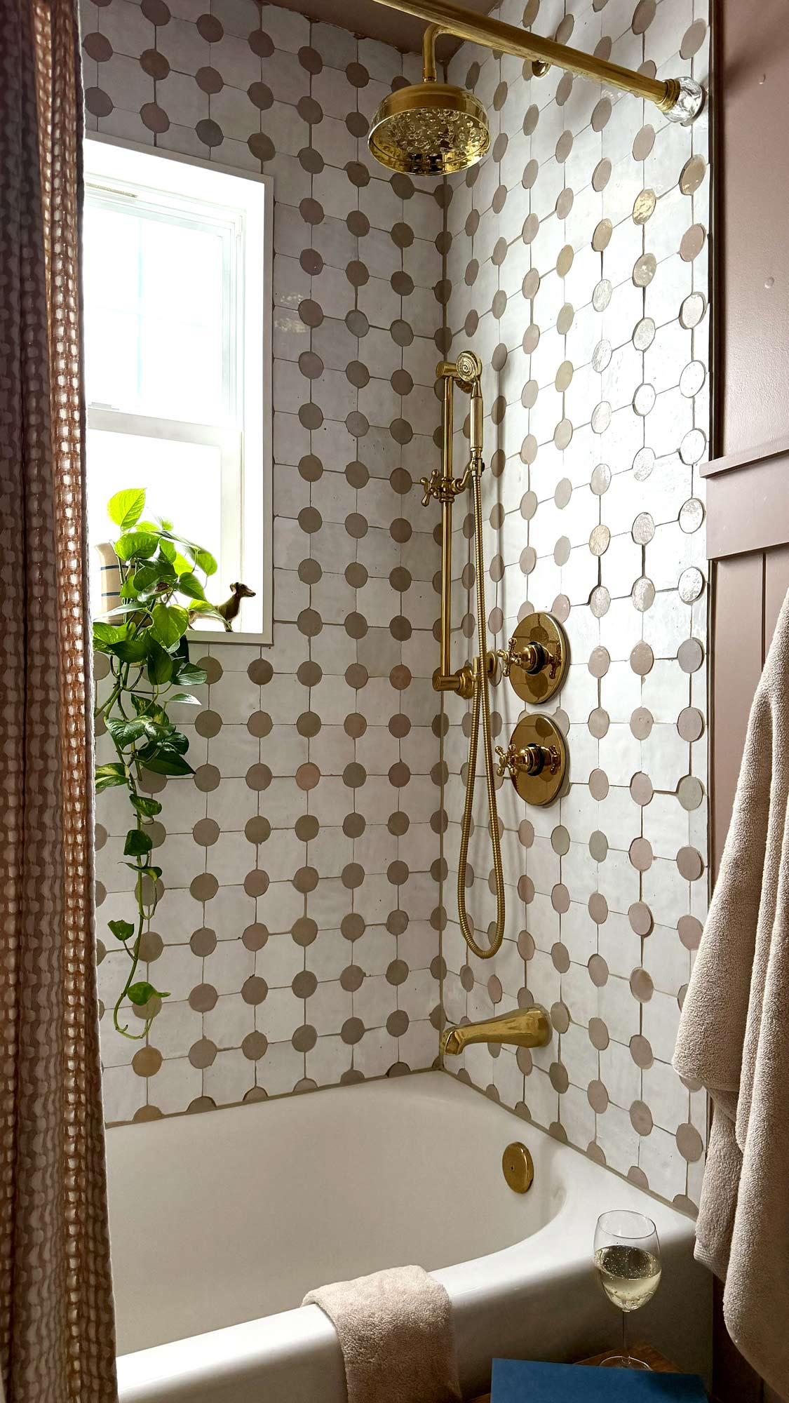 Bathtub with gold fixtures, patterned tile walls, hanging plant by window, towel, and glass of white wine.