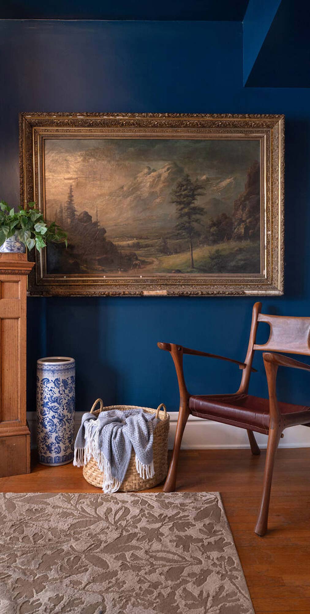 Room corner with large landscape painting, wooden chair, wicker basket with blanket, and blue vase.