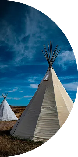 Two traditional tipis on a grassy plain under a bright blue sky with wispy clouds.