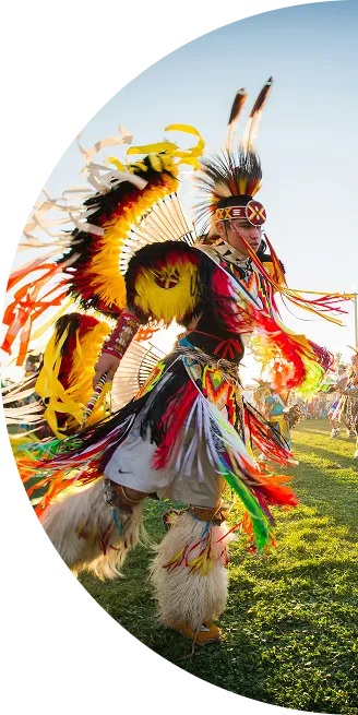 Native American dancer in colorful traditional regalia performing at outdoor event on grass under blue sky.