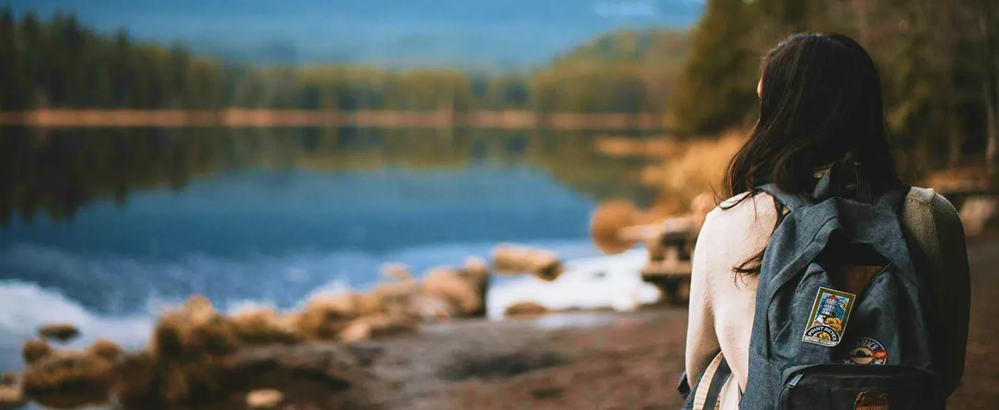 Person with backpack sitting on a rock by a lake surrounded by trees and mountains in the distance.