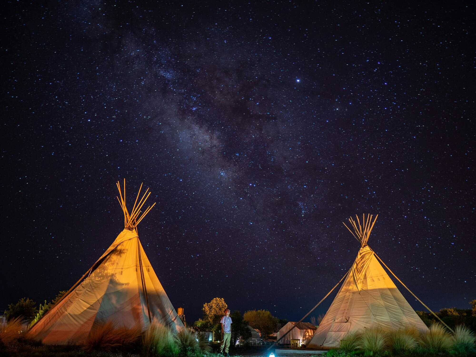 Two lit teepees at night with a star-filled sky and a person standing between them.