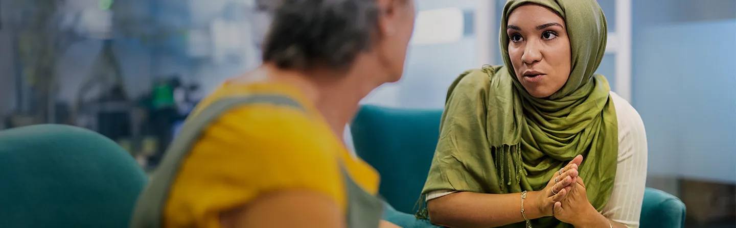 Woman in a green hijab attentively listening and talking to a person in a yellow shirt indoors.
